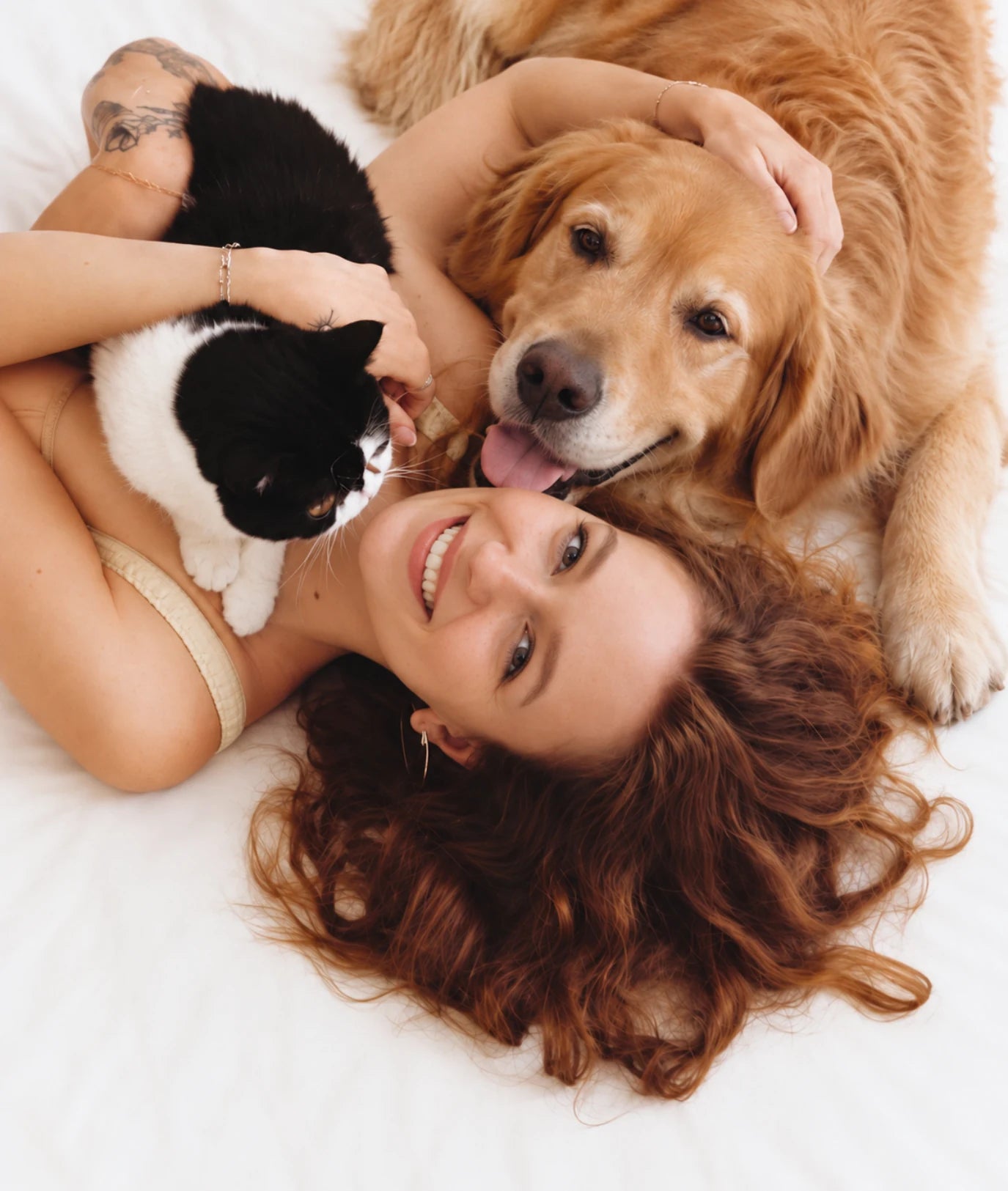 A woman lying on white linen with her cat and golden retriever