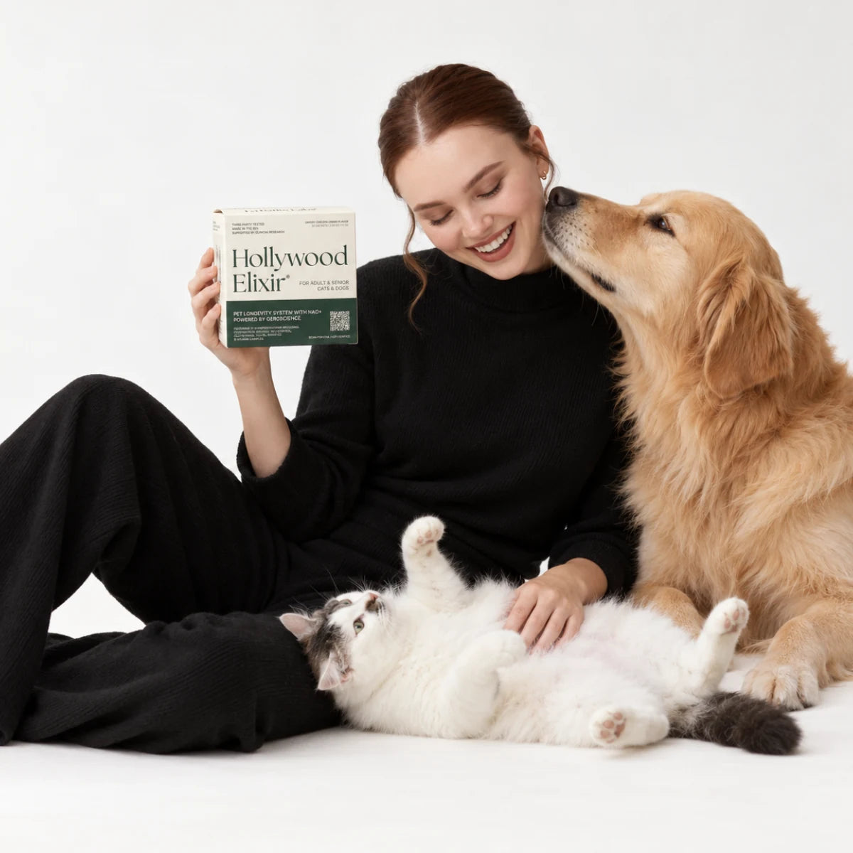 A woman holding Hollywood Elixir beside a golden retriever and a reclining cat