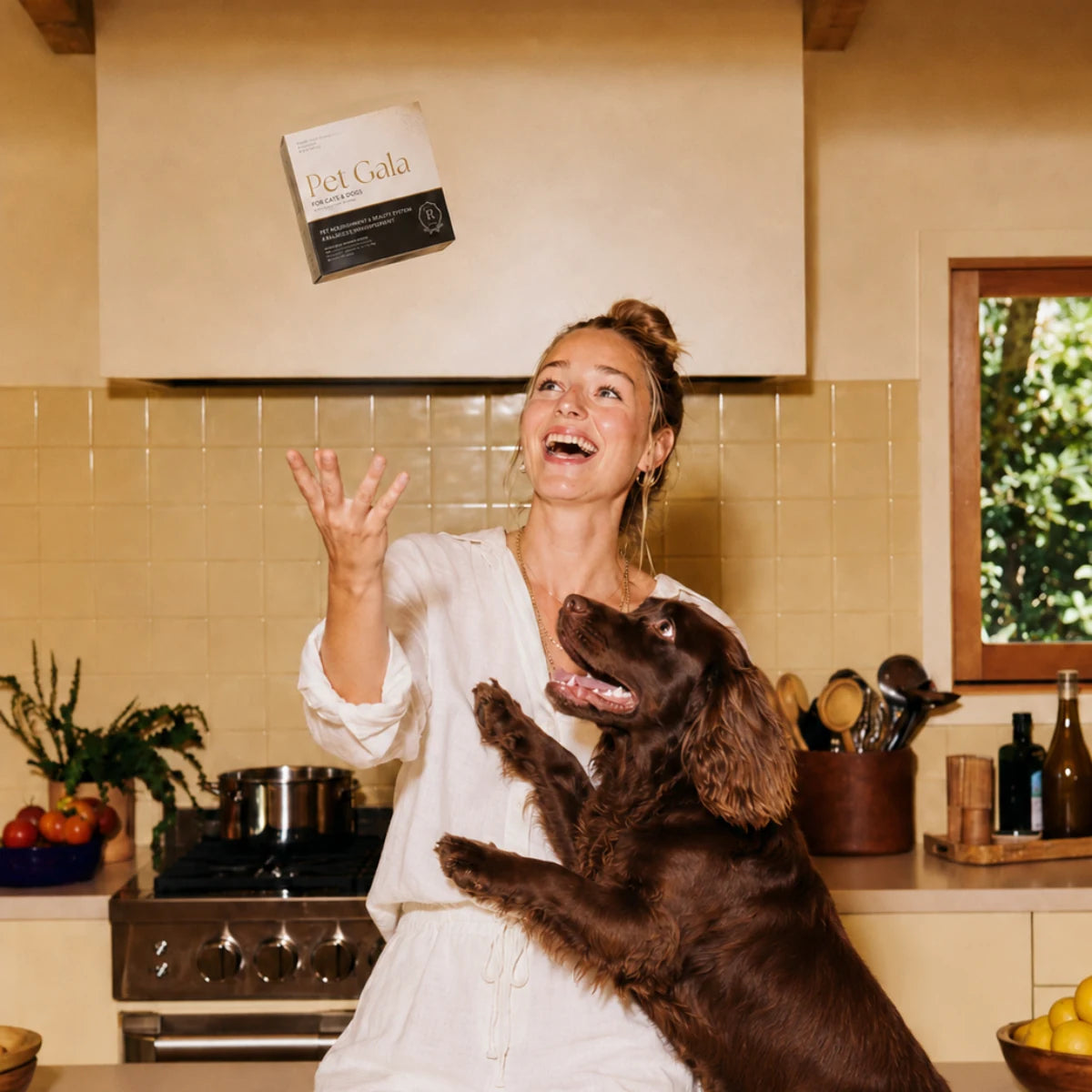 A woman tossing a Pet Gala box beside her brown dog in a kitchen