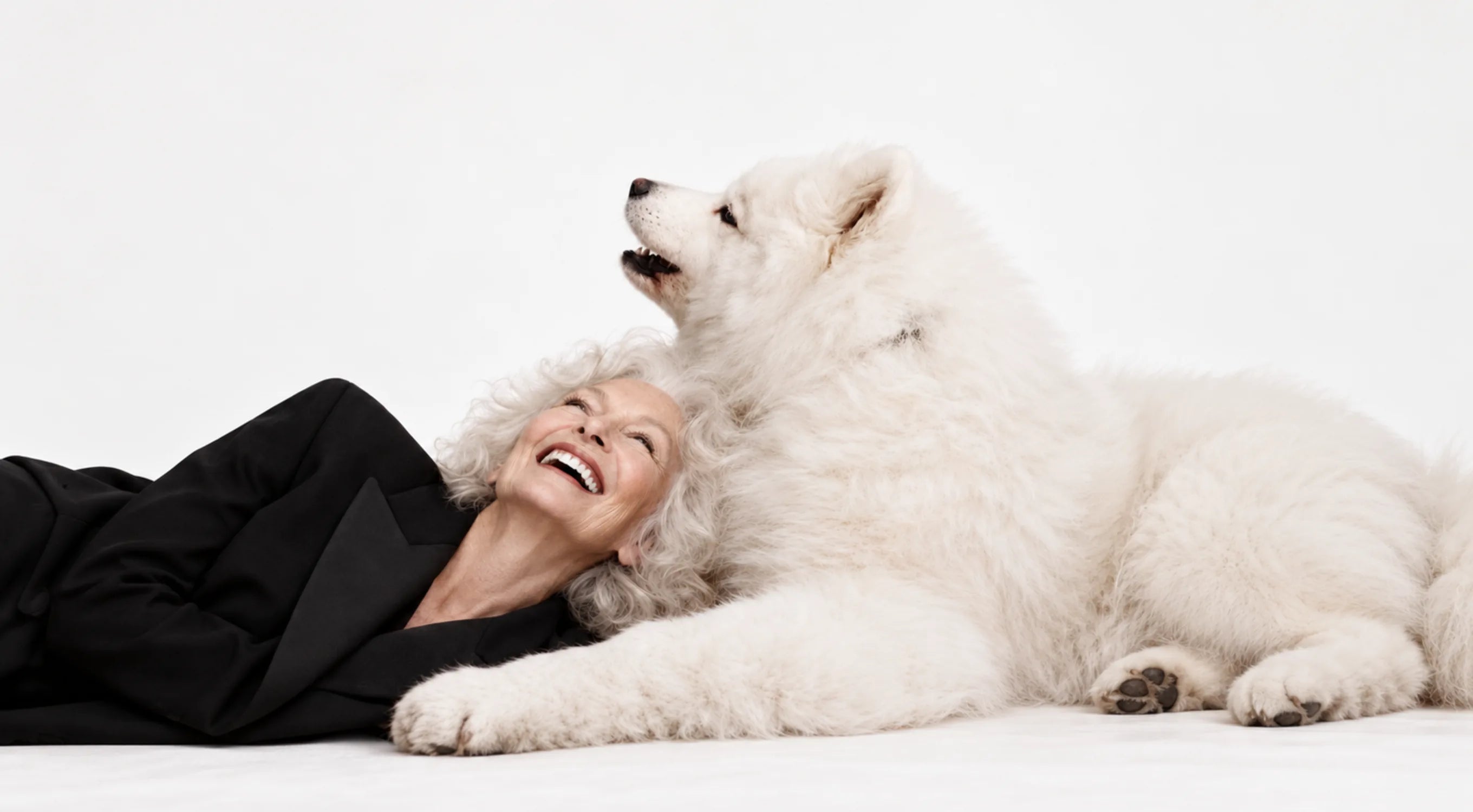 A laughing older woman lying on the floor beside her white Samoyed